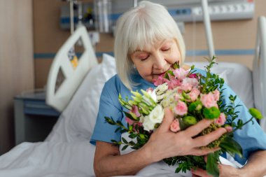 selective focus of recovering senior woman on bed with flowers in hospital clipart