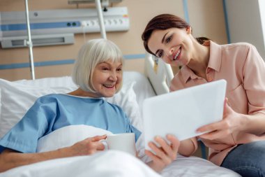 happy senior woman and daughter using digital tablet in hospital bed