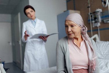 upset senior woman in kerchief with cancer sitting on hospital bed with female doctor holding diagnosis on background clipart