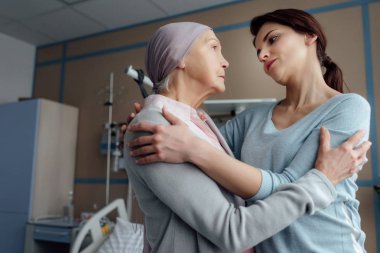 selective focus of worried daughter hugging sick senior mother with cancer in hospital clipart