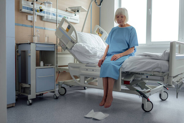 upset lonely senior woman sitting on bed in hospital ward 