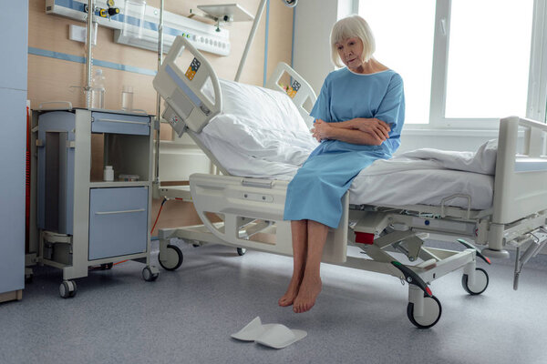 sad senior woman with arms crossed sitting on bed in hospital ward 