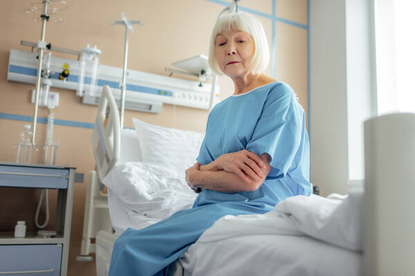 lonely senior woman with arms crossed sitting on bed in hospital ward 