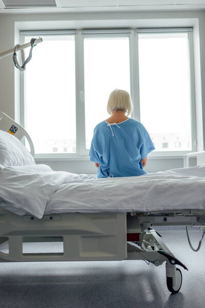 back view of lonely senior woman sitting on bed in hospital ward 