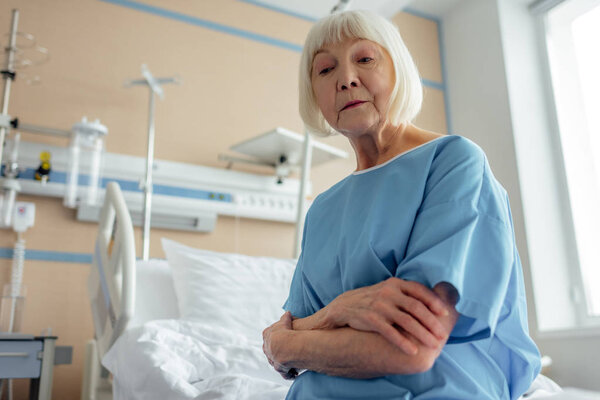 selective focus of sad senior woman with arms crossed sitting on bed in hospital ward 