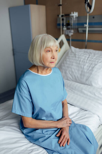 sad senior woman with folded hands sitting on bed in hospital