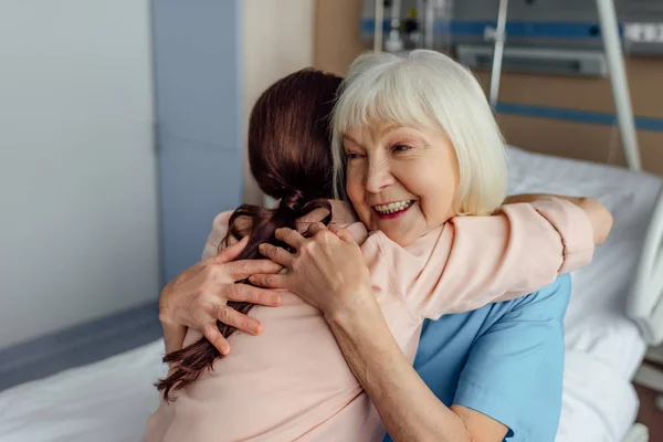 开朗的老年妇女和女儿坐在床上 在医院拥抱 — 图库照片