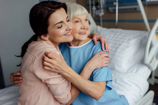 Lächelnde Seniorin Und Tochter Sitzen Auf Bett Und Umarmen Sich — Stockfoto