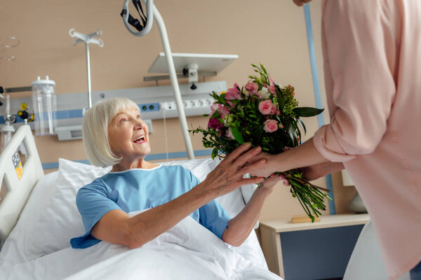 daughter presenting flowers to smiling senior woman lying in bed in hospital