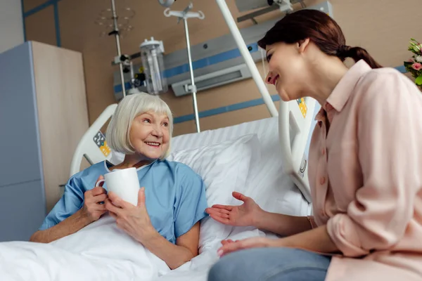 老年妇女躺在床上喝茶 同时与女儿在医院交谈 — 图库照片