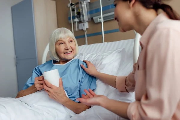 happy senior woman lying in bed and drinking tea while talking to daughter in hospital