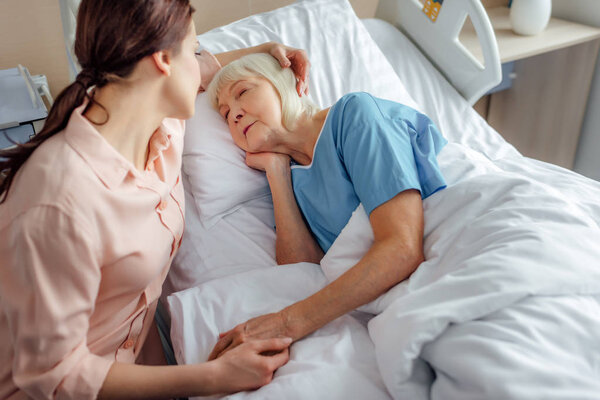 selective focus of daughter sitting near senior mother in bed and holding hands in hospital