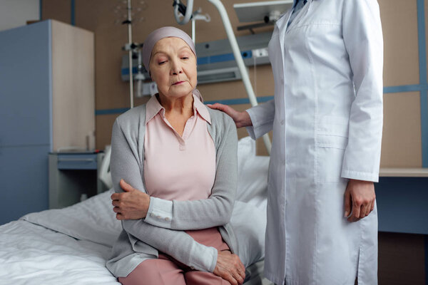 female doctor consoling upset senior woman with cancer and arms crossed in hospital