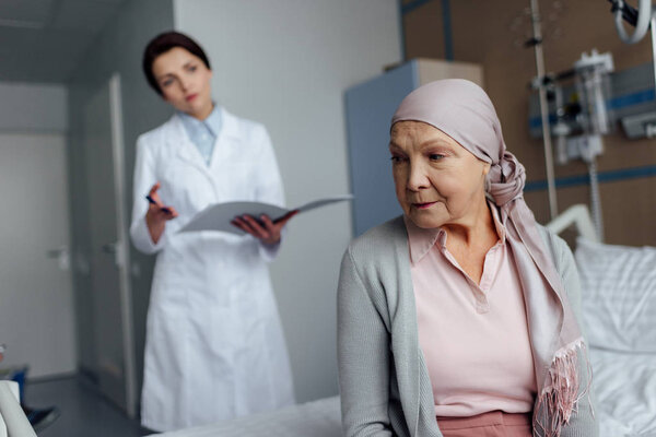 upset senior woman in kerchief with cancer sitting on hospital bed with female doctor holding diagnosis on background