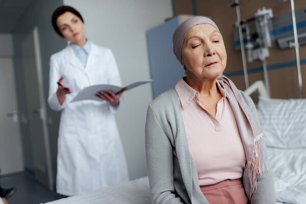 sad senior woman in kerchief with cancer sitting on hospital bed with female doctor holding diagnosis on background