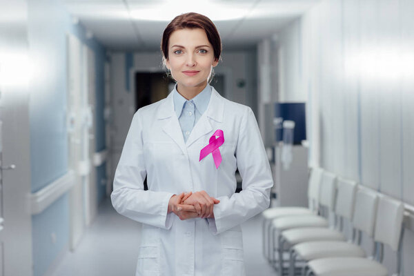 female doctor with pink ribbon and folded hands looking at camera in hospital, breast cancer awareness concept