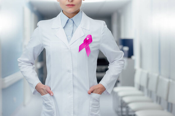 cropped view of female doctor with pink ribbon and hands akimbo in hospital, breast cancer awareness concept