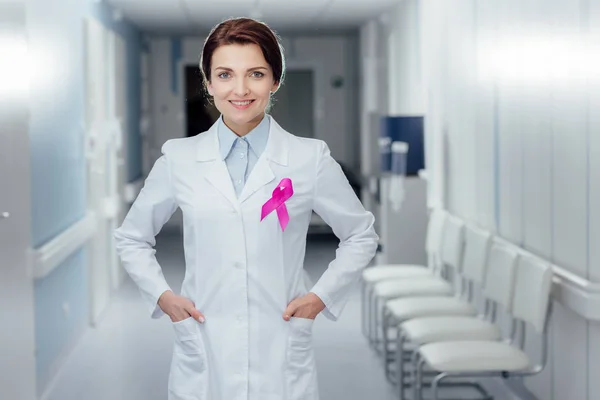 smiling female doctor with pink ribbon and hands akimbo in hospital, breast cancer awareness concept