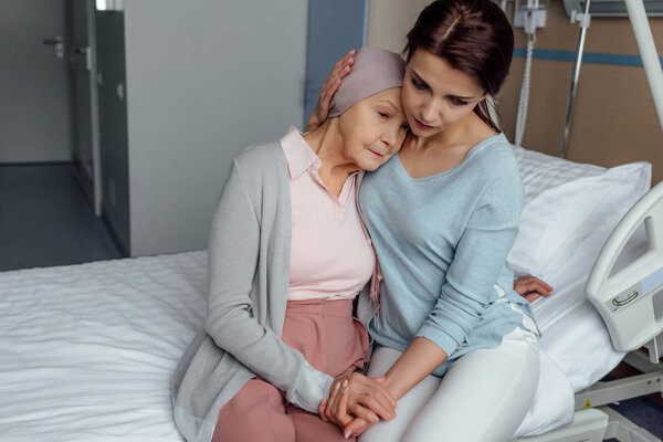 worried daughter hugging senior sad mother with cancer in hospital