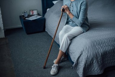 partial view of senior woman sitting on bed with walking stick at home clipart