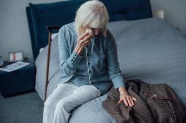 crying senior woman sitting on bed, wiping tears and looking at jacket at home clipart