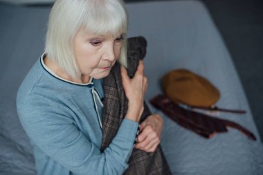 upset senior woman with grey hair sitting on bed and holding jacket at home clipart