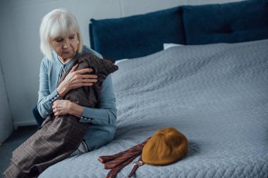 upset senior woman sitting on bed and holding jacket at home clipart