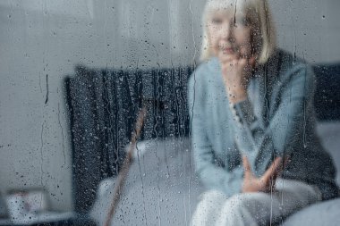 lonely senior woman sitting on bed and propping chin with hand at home through window with raindrops clipart