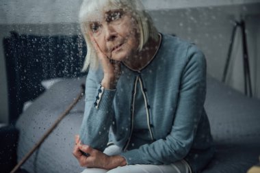 sad senior woman sitting on bed at home through window with raindrops clipart