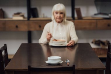 selective focus of bowl and spoon on table with senior woman eating on background clipart