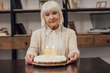 Evde Doğum günü pastası ile masada oturan gri saçlı üzgün üst düzey kadın 
