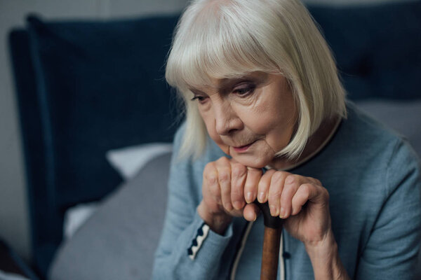 portrait of upset senior woman with hands on walking stick sitting on bed at home