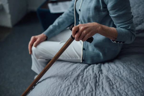 裁剪视图的高级妇女坐在床上 拿着拐杖在家里 — 图库照片