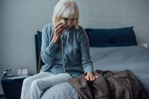 selective focus of crying senior woman sitting on bed, wiping tears and looking at jacket at home