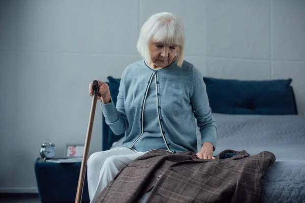 悲伤的老年妇女坐在床上 拿着拐杖 看着夹克在家里 — 图库照片