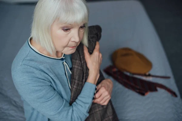 心烦意乱的老太婆与灰色的头发坐在床上 并在家里举行夹克 — 图库照片