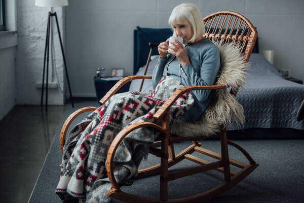 senior woman sitting in wicker rocking chair with blanket and drinking coffee at home