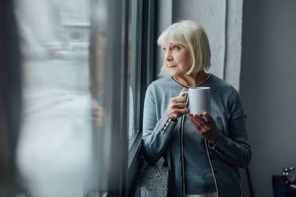 Задумчивая Пожилая Женщина Держит Чашку Кофе Смотрит Окно Дома — стоковое фото