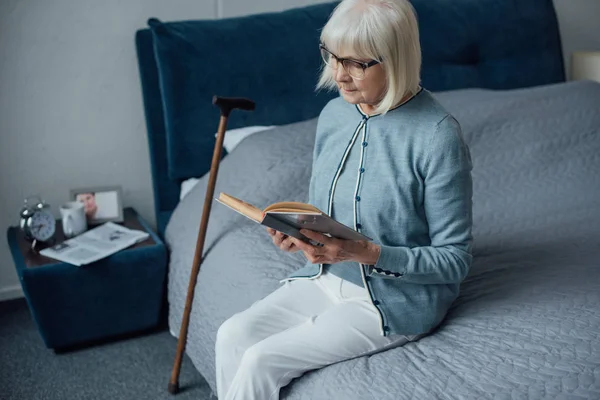 Nachdenkliche Seniorin Mit Brille Sitzt Auf Bett Und Liest Buch — Stockfoto