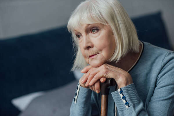 portrait of thoughtful senior woman with hands on walking stick sitting on bed at home