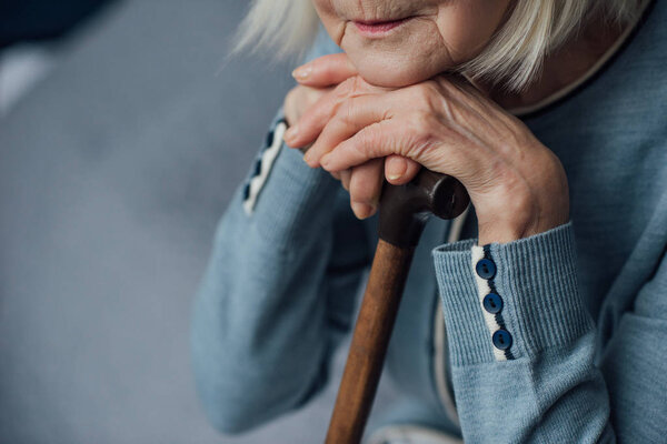cropped view of senior woman with hands on walking stick sitting on bed at home