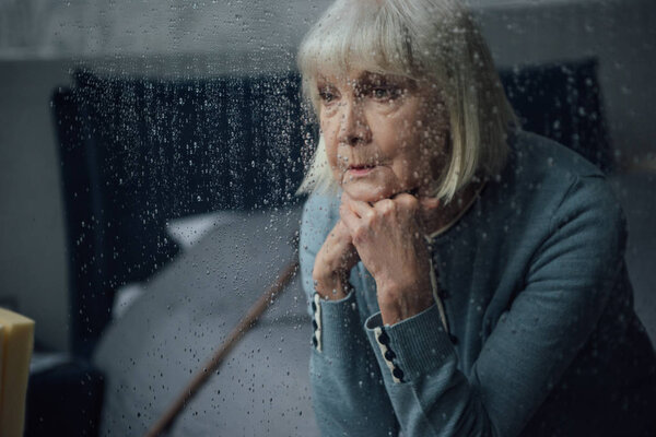 upset senior woman sitting and propping chin with hands at home through window with raindrops