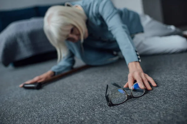 孤独的高级女子躺在地板上 感觉身体不适 并在家里伸手去拿眼镜 — 图库照片
