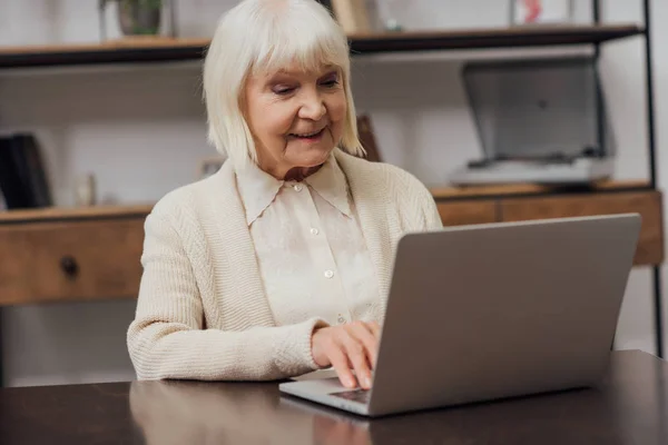 快乐的老年妇女坐在桌子上 在家里的笔记本电脑上打字 — 图库照片