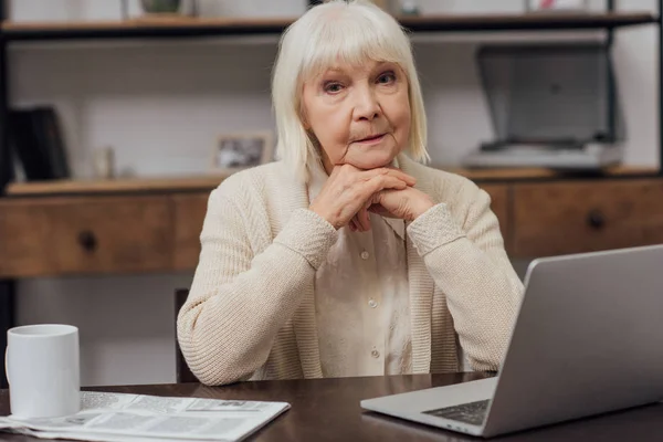 坐在桌旁的老年妇女拿着笔记本电脑 看着相机 双手拿着手在家里支撑着下巴 — 图库照片