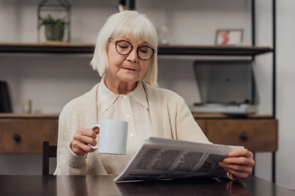 戴眼镜的高级妇女坐在桌旁 在家里喝咖啡 看报纸 — 图库照片