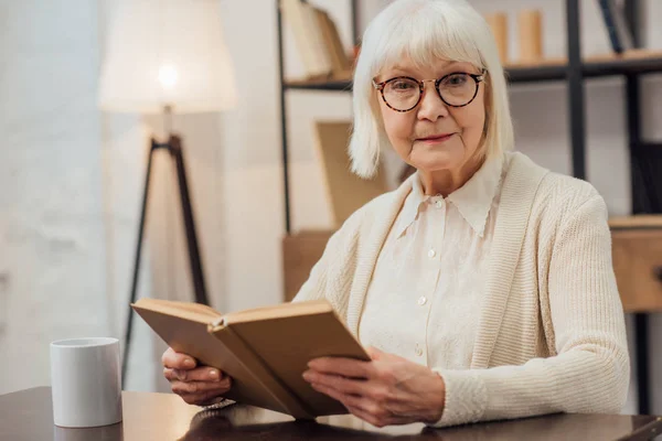 Senior Woman Glasses Sitting Table Looking Camera While Reading Book — Stock Photo, Image