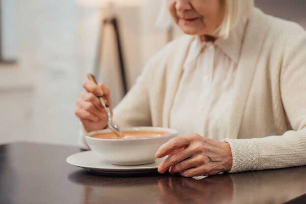 cropped view of senior woman sitting and eating cream soup at home