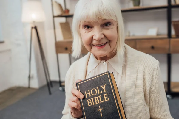 Улыбающаяся Пожилая Женщина Смотрит Камеру Держит Священную Библию Дома — стоковое фото