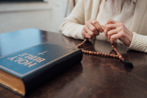 partial view of senior woman sitting at table with rosary and holy bible at home